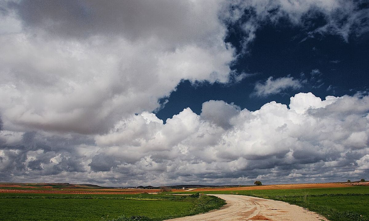 Camino de tierra entre campos verdes de la campiña manchega bajo grandes nubes